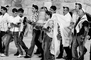 A procession in the Old City of Jerusalem escorting a Bar Mitzvah boy with singing to the Kotel (Western Wall) where he will be called to read from the Torah for the first time. Note that the participants are wearing tefillin which indicates that this is a weekday procession. Casual dress is typical of Israelis even for events such as a Bar Mitzvah.