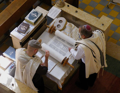A Torah scroll being read in synagogue. As the text of a Torah scroll is written without vowels, a second man stands adjacent to the reader and checks his reading against a printed edition with vowels, thus enabling him to quickly call out a corretion to any error made by the reader